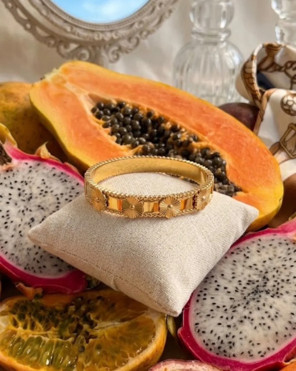 Bracelet Lila ton or à motifs floraux, posé sur un coussin de lin parmi des fruits coupés